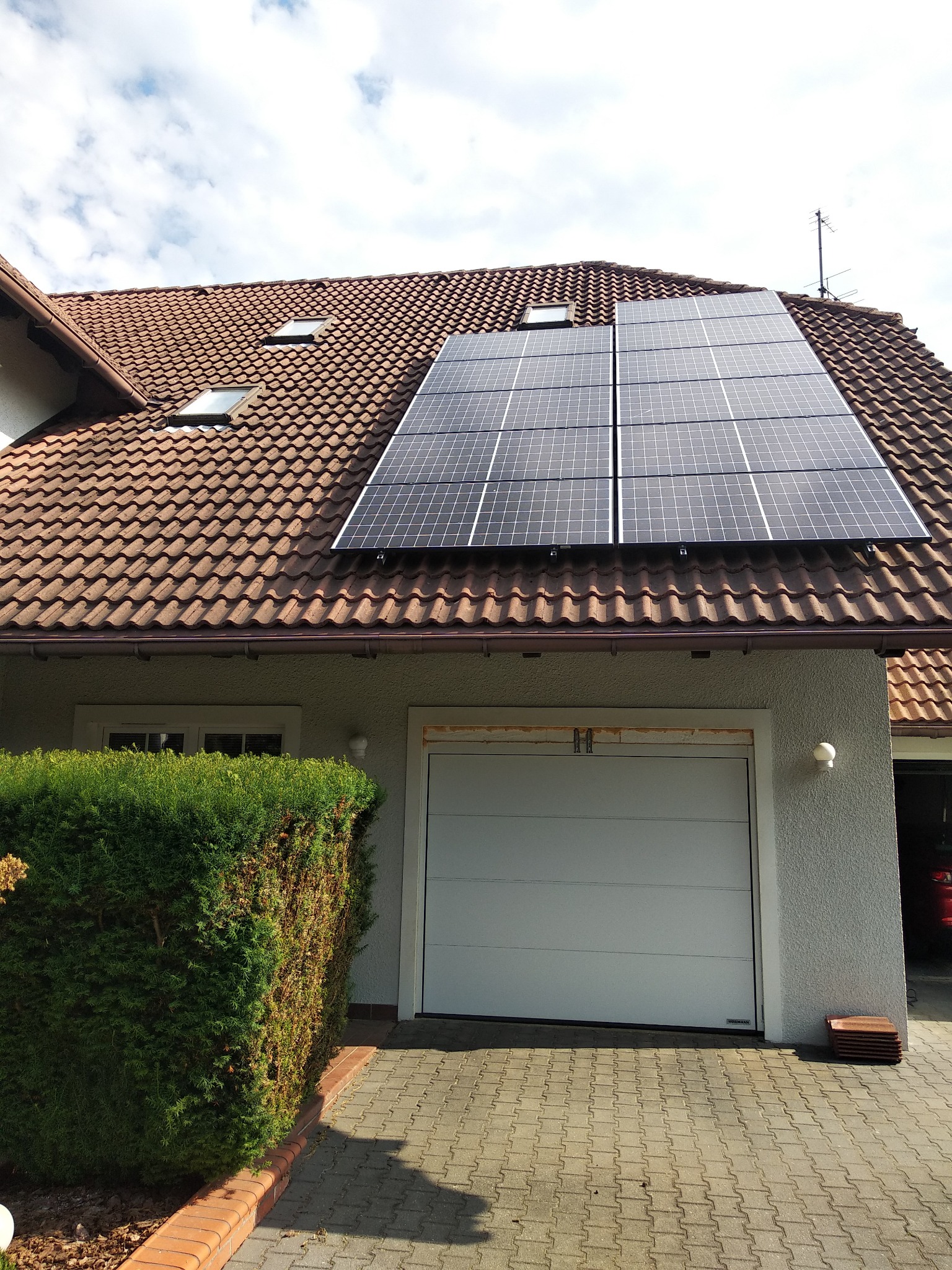 Cztery panele słoneczne zamontowane na brązowym dachu z dachówki ceramicznej domu jednorodzinnego z garażem, widoczne okna dachowe, antena telewizyjna i starannie przycięty żywopłot.