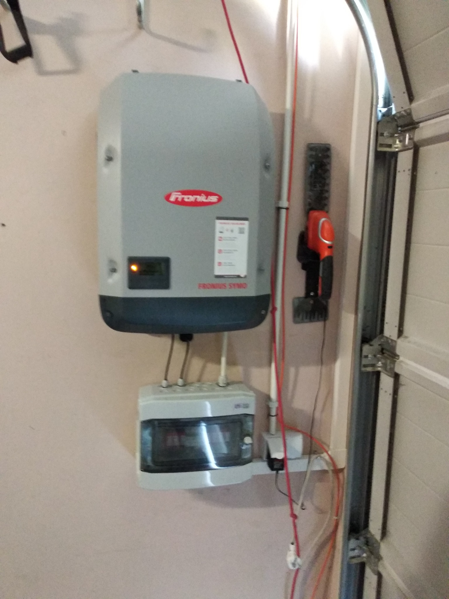 Szary falownik Fronius zamontowany na jasnoróżowej ścianie garażu, z widocznymi przewodami i skrzynką przyłączeniową pod spodem, obok wiszą elektryczne nożyce do żywopłotu.