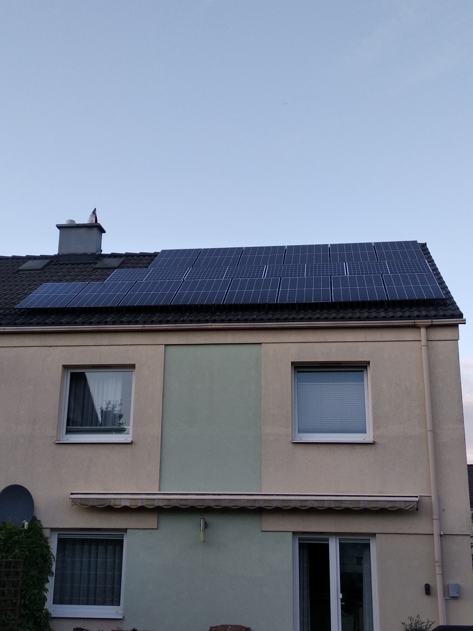 Dwukondygnacyjny dom z beżową elewacją i panelami fotowoltaicznymi na ciemnym dachu, widoczne okna z roletami, markiza nad oknem na parterze oraz antena satelitarna po lewej stronie.