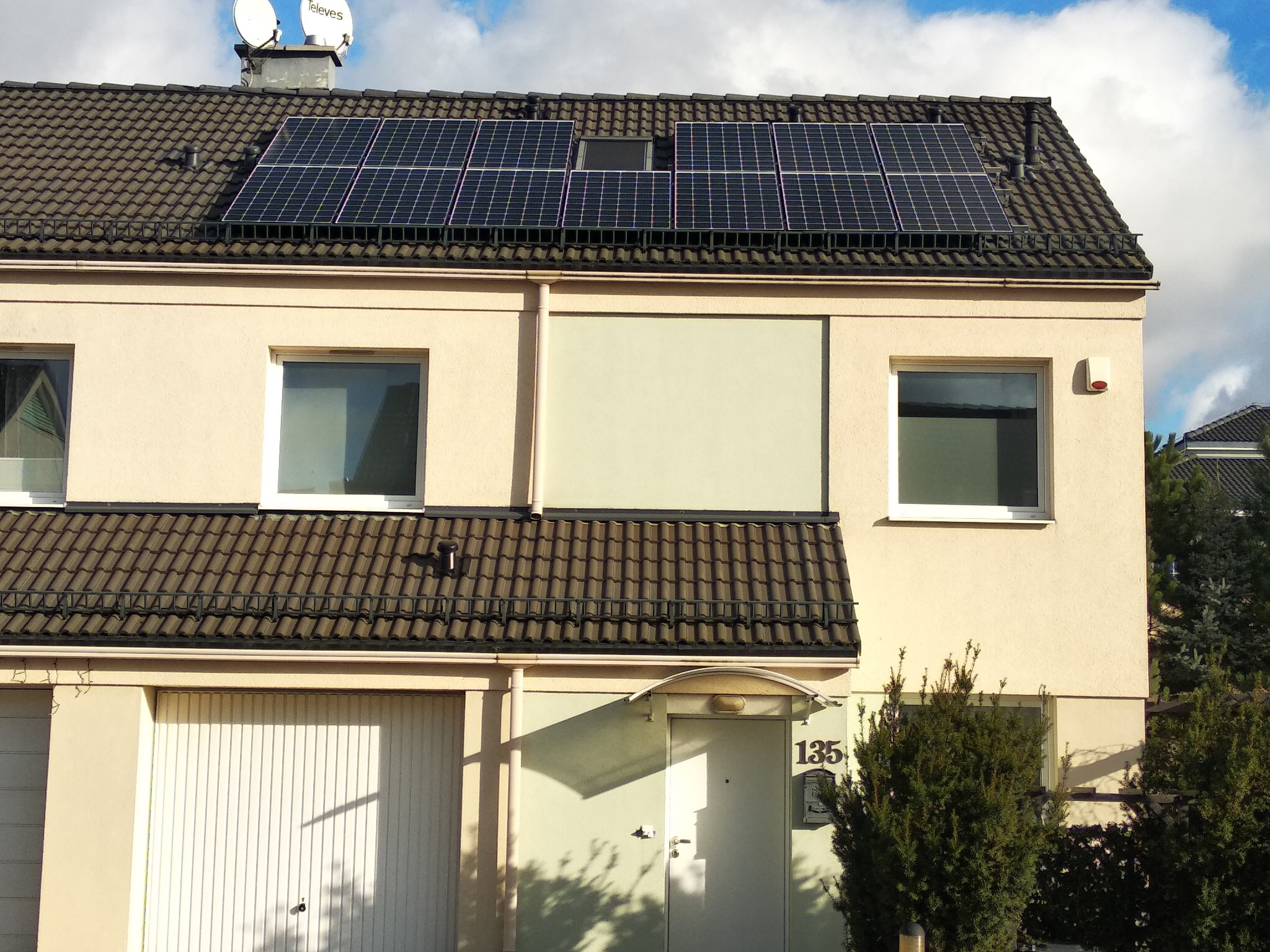 Panele słoneczne na dachu dwupiętrowego domu szeregowego z brązową dachówką, widoczne anteny satelitarne oraz numer domu 135 nad wejściem.