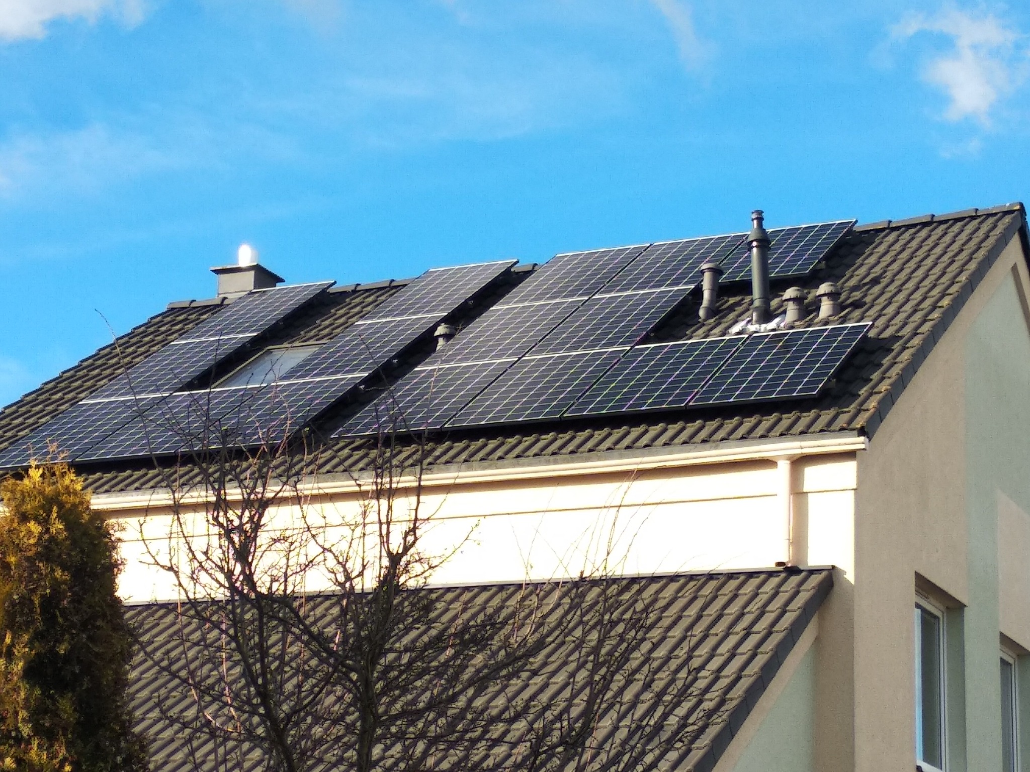 Panele słoneczne na dachu domu jednorodzinnego z brązową dachówką, widoczne kominy i drzewo na pierwszym planie, błękitne niebo w tle.