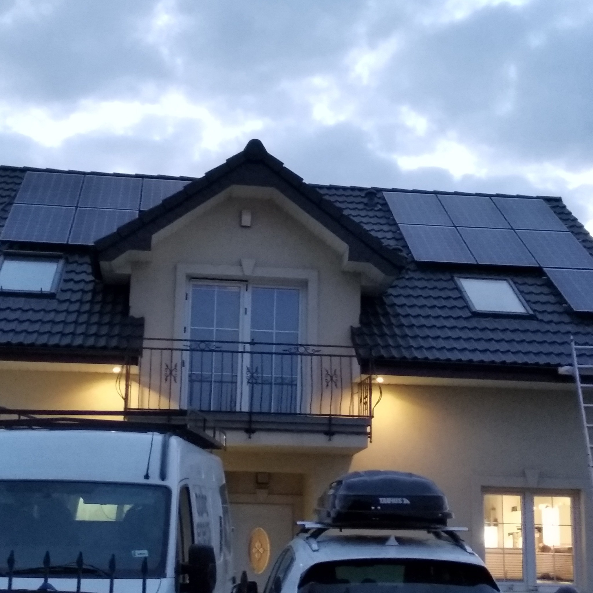 Dom jednorodzinny z zainstalowanymi panelami fotowoltaicznymi na dachu, widoczne okna dachowe, balkon z ozdobną balustradą, samochód osobowy z bagażnikiem dachowym i częściowo widoczny biały bus.