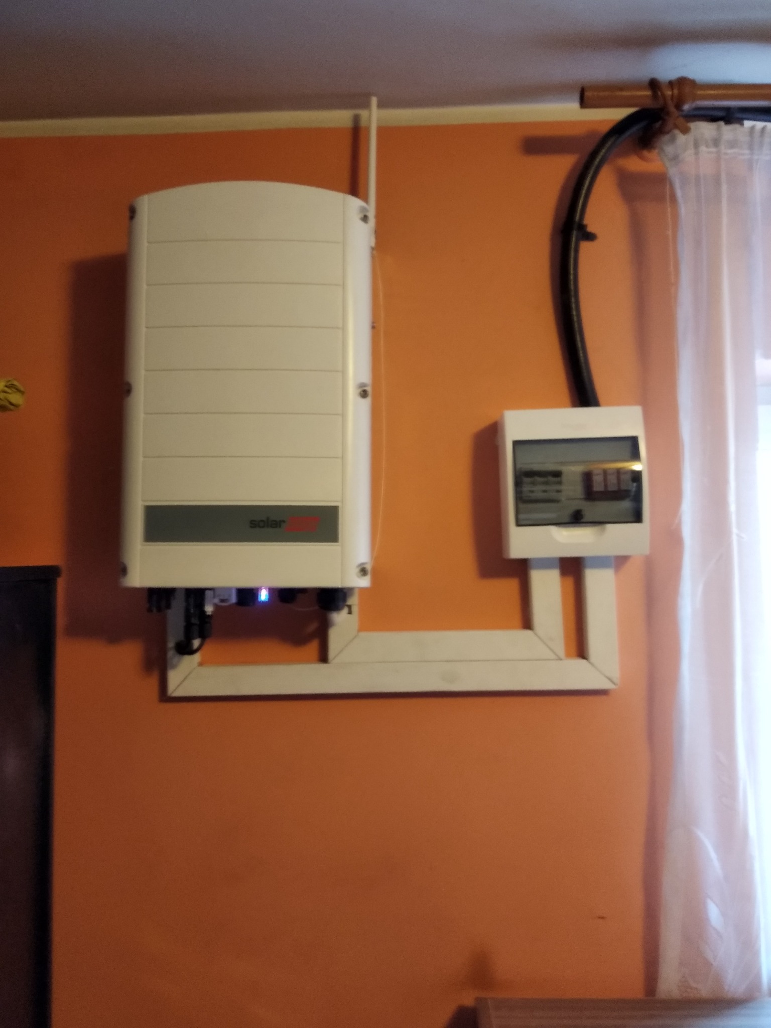 Biały inwerter z logo SolarEdge zamontowany na pomarańczowej ścianie obok skrzynki elektrycznej z widocznymi bezpiecznikami, z kablem biegnącym wzdłuż ściany do skrzynki, widoczny fragment zasłony...