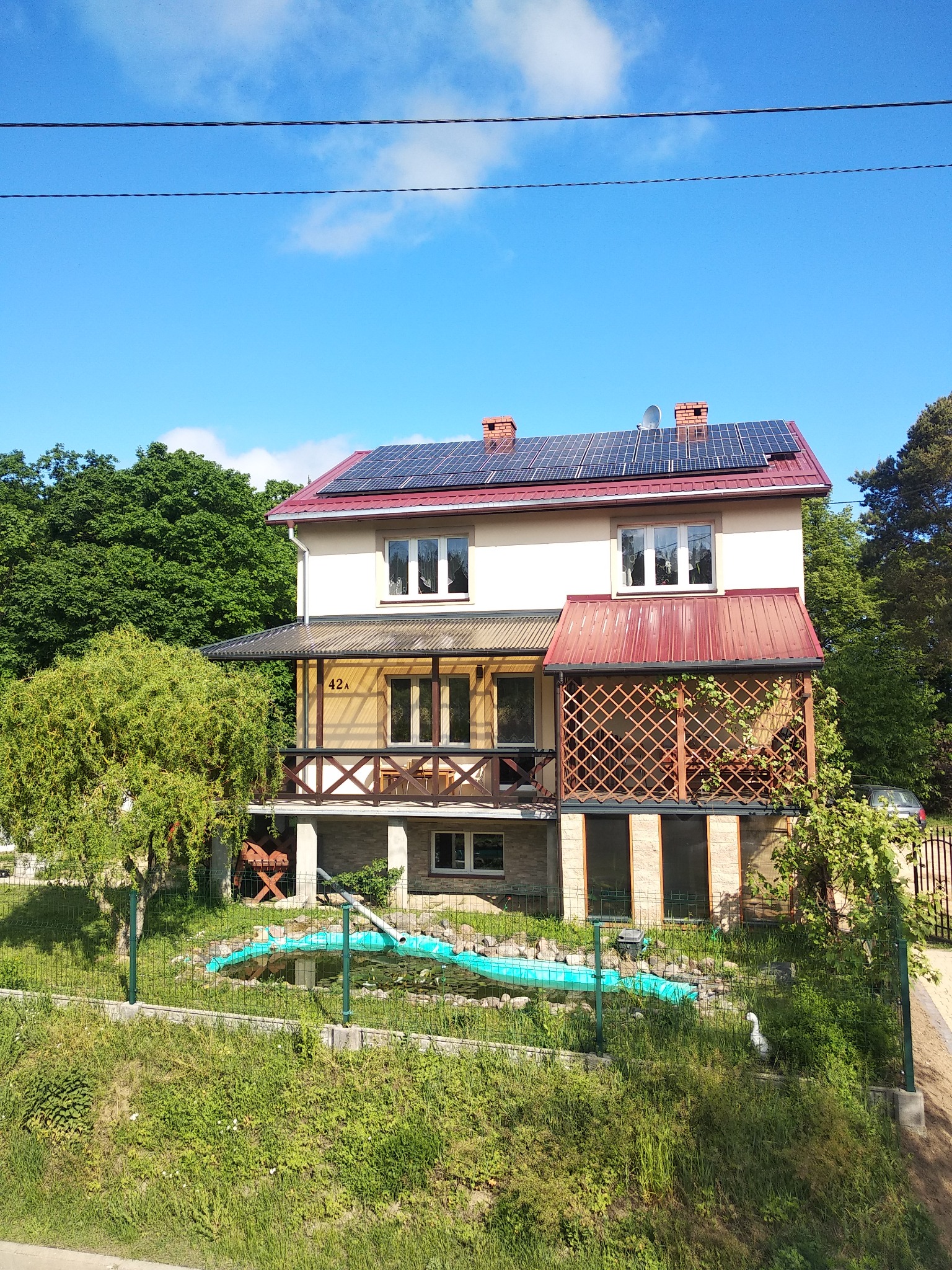 Dwukondygnacyjny dom z panelami słonecznymi na czerwonym dachu, widok z góry na ogród z oczkiem wodnym i dekoracyjnym ogrodzeniem.