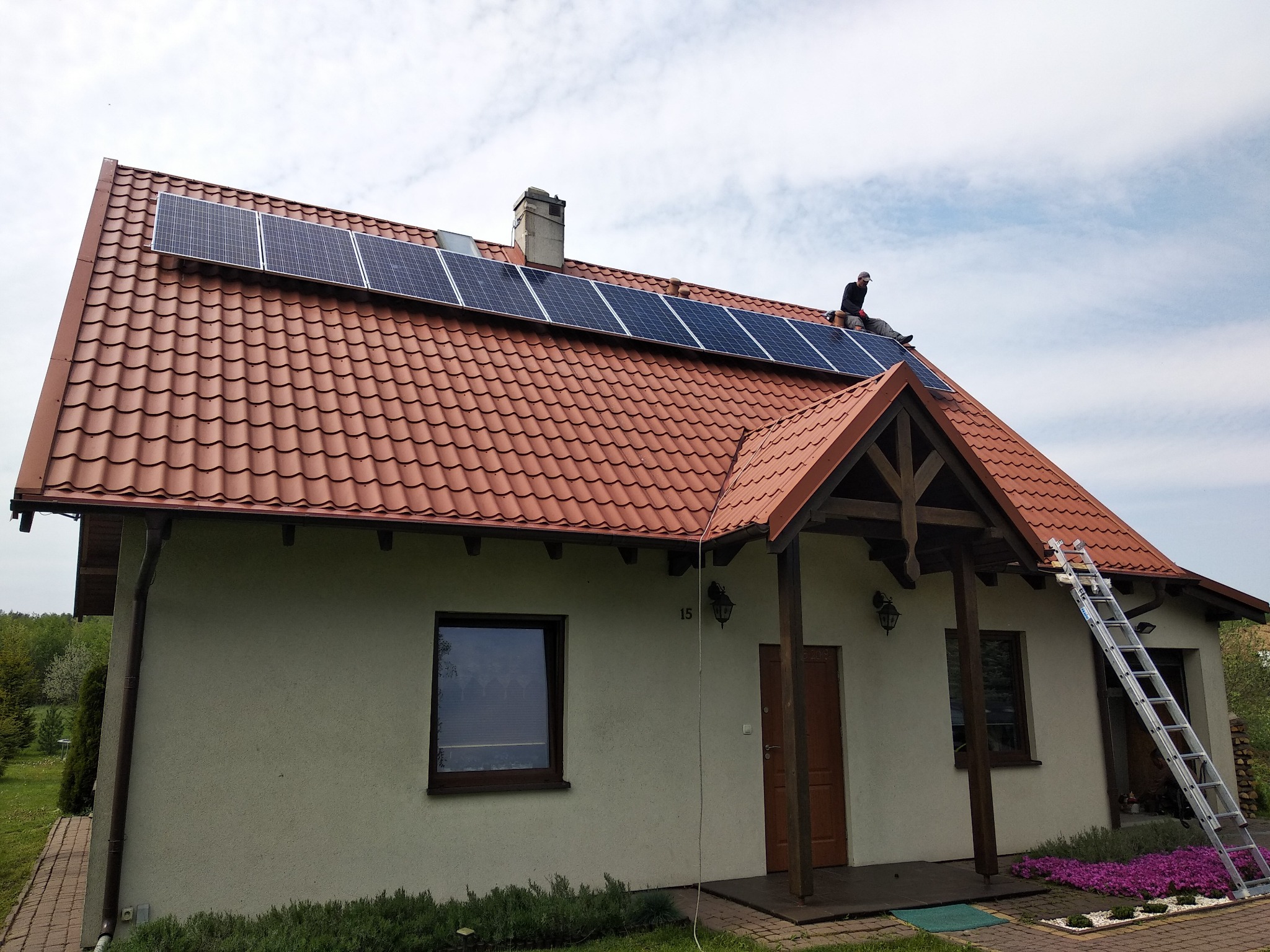 Montaż paneli fotowoltaicznych na dachu domu z brązową dachówką, widoczna drabina i instalator w czapce podczas pracy.