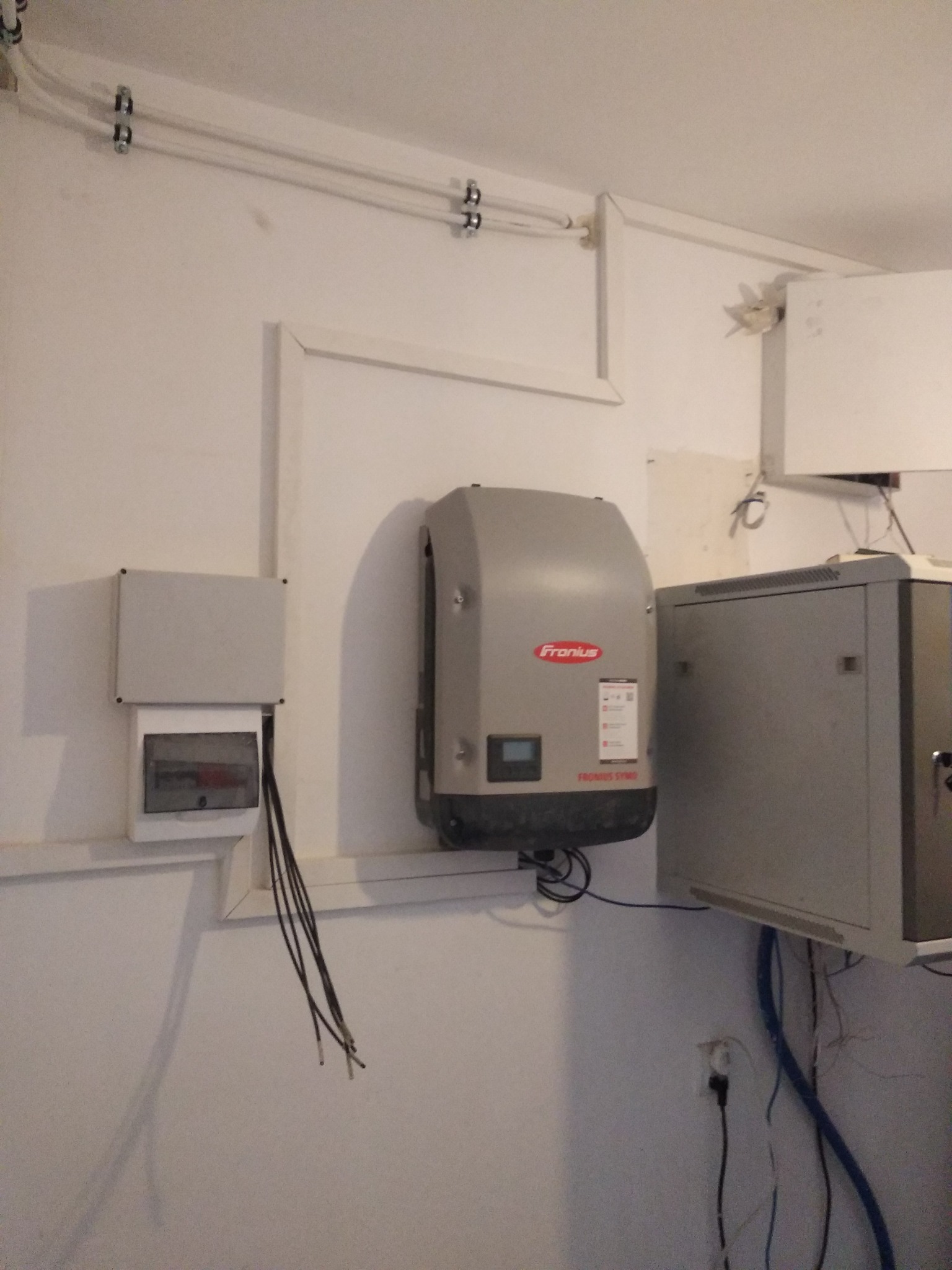 Szary inwerter Fronius Symo zamontowany na białej ścianie, obok skrzynka elektryczna z wystającymi kablami i szara szafa sterownicza. Widoczne rurki instalacyjne pod sufitem.