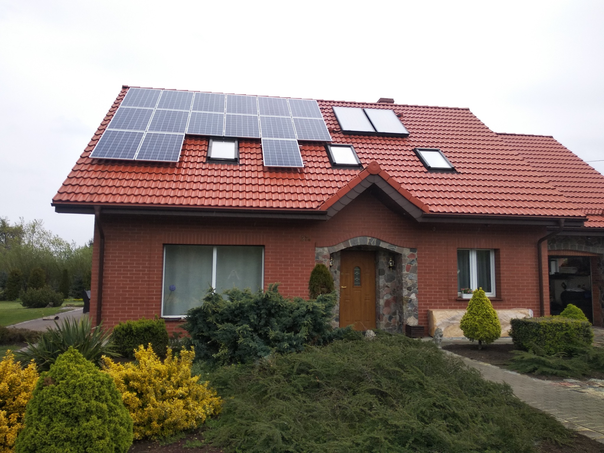 Dom jednorodzinny z czerwoną dachówką i panelami słonecznymi umieszczonymi na dachu obok okien dachowych, widok z poziomu ogrodu z różnorodną roślinnością.