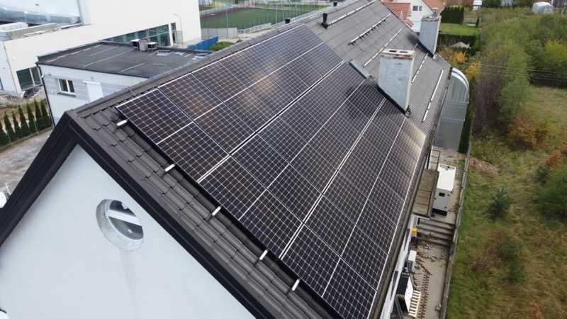 Panele słoneczne na dachu domu z ciemnej dachówki, widok z góry, z widocznym kominem i oknem w kształcie okręgu na ścianie budynku.