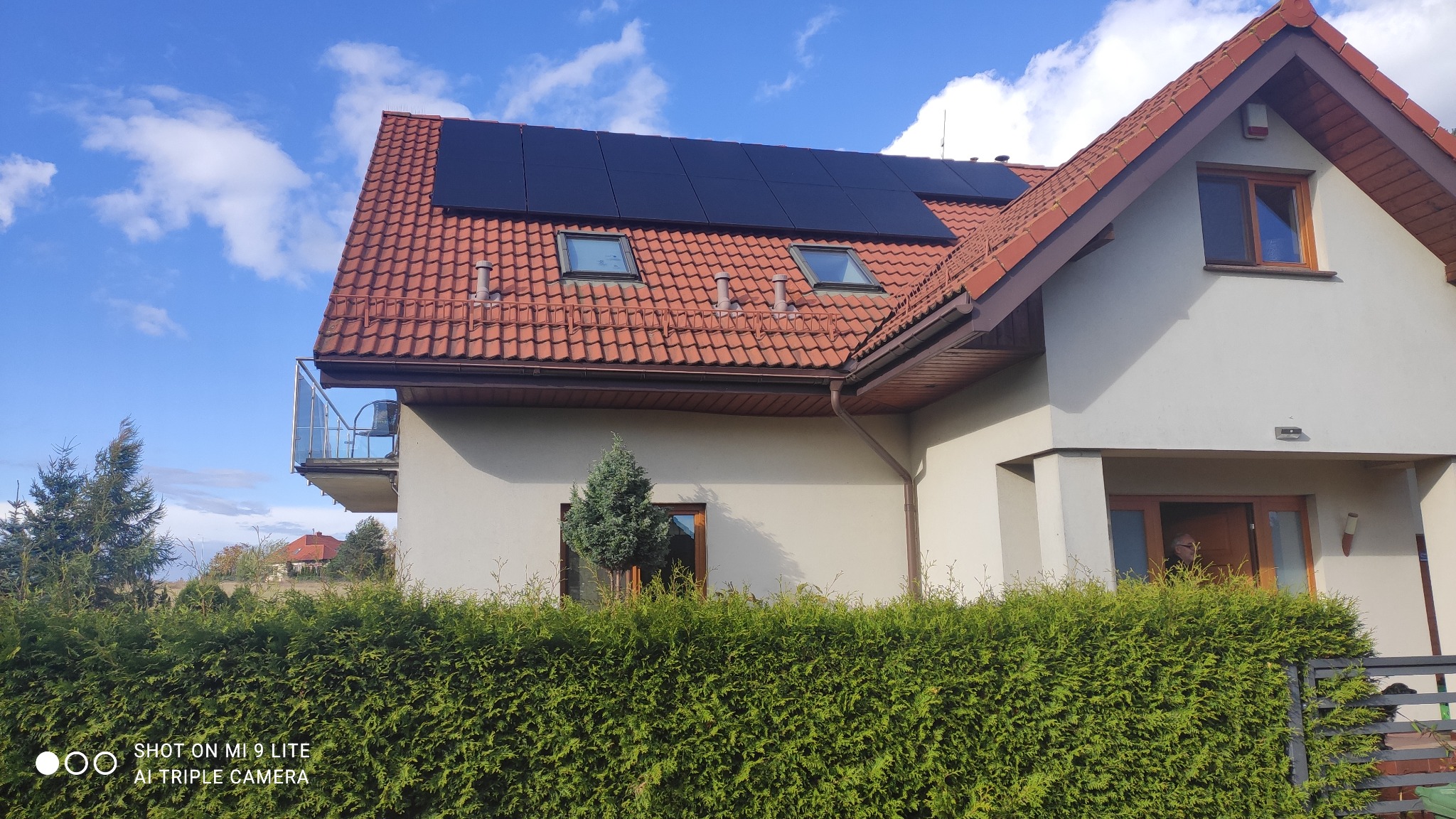 Dom jednorodzinny z ciemnymi panelami słonecznymi na dachu z czerwonej dachówki, widok zza żywopłotu w słoneczny dzień.