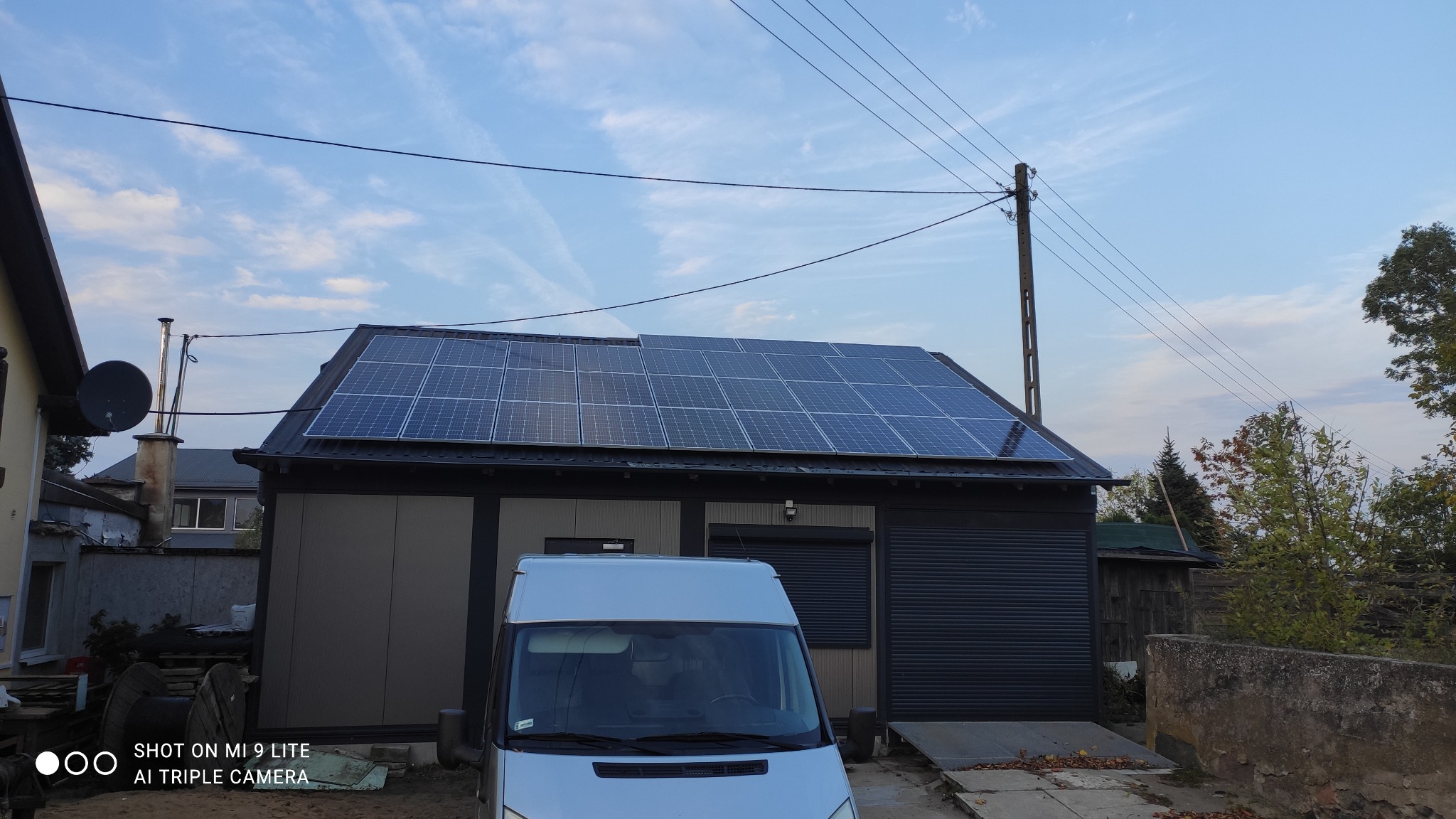 Panele słoneczne na dachu budynku gospodarczego, widok z poziomu ulicy, z zaparkowanym białym vanem na pierwszym planie.