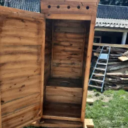 Surowa, drewniana toaleta zewnętrzna z otwartymi drzwiami, widoczna na trawniku, z drabiną opartą o stertę drewna w tle.