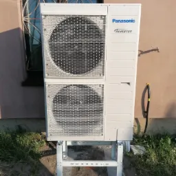 Zewnętrzna jednostka klimatyzacji Panasonic Inverter zamontowana na metalowym stelażu przy ścianie budynku z oknem zabezpieczonym ozdobną kratą.