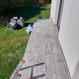 Krzysiek - Ułożona szara podłoga z płytek imitujących drewno na balkonie, widok z góry, drabina oparta o podłogę, w tle trawa, zaparkowany srebrny bus i materiały budowlane.