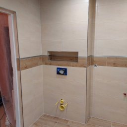 Krzysiek - Wnętrze łazienki w trakcie prac wykończeniowych, widoczne beżowe płytki na ścianach z dekoracyjnym pasem imitującym drewno, płytki podłogowe w odcieniach drewna, zamontowany stelaż podtynkowy WC...