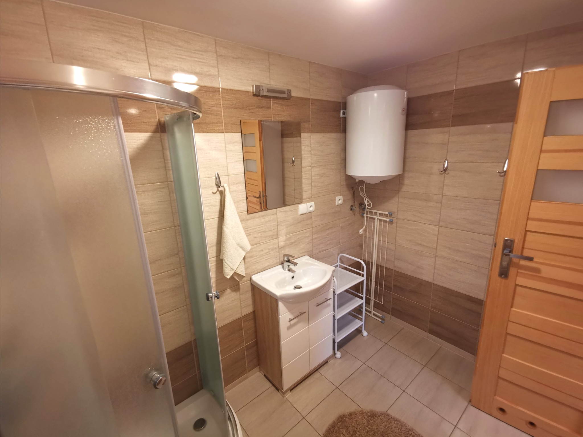 Wykończona łazienka z kabiną prysznicową z półokrągłymi drzwiami, umywalką z szafką, grzejnikiem elektrycznym, lustrem i drewnianymi drzwiami.