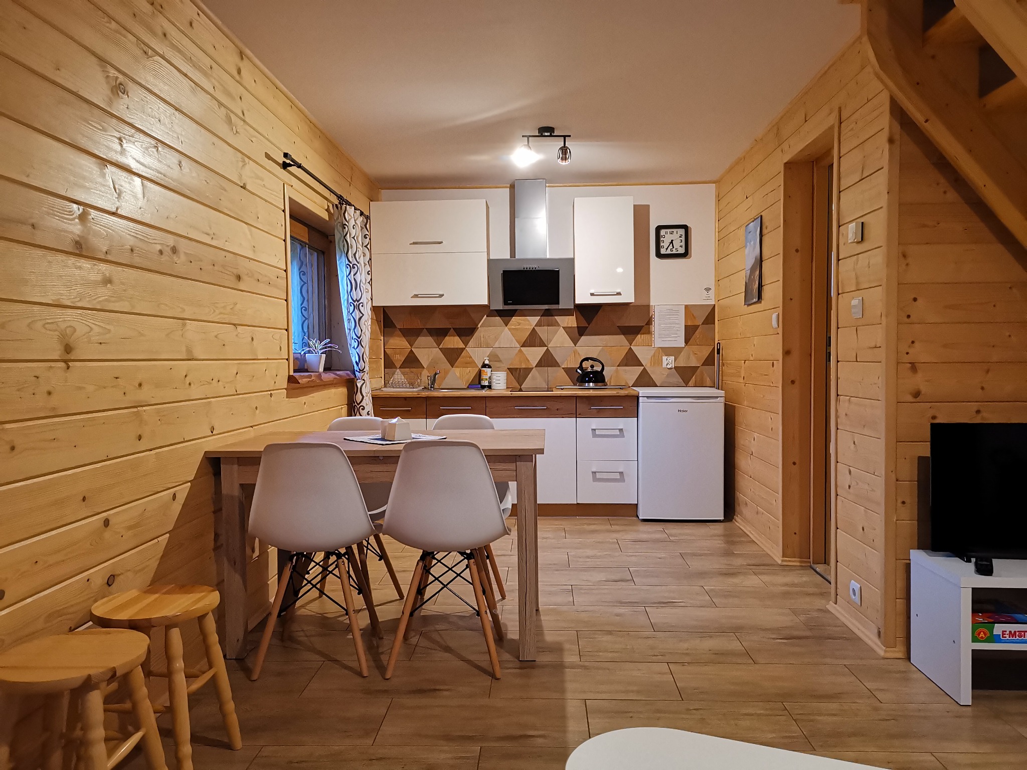 Drewniane wnętrze domu z jadalnią i aneksem kuchennym, widoczne krzesła Eames, stół, hokery, białe szafki kuchenne i lodówka.