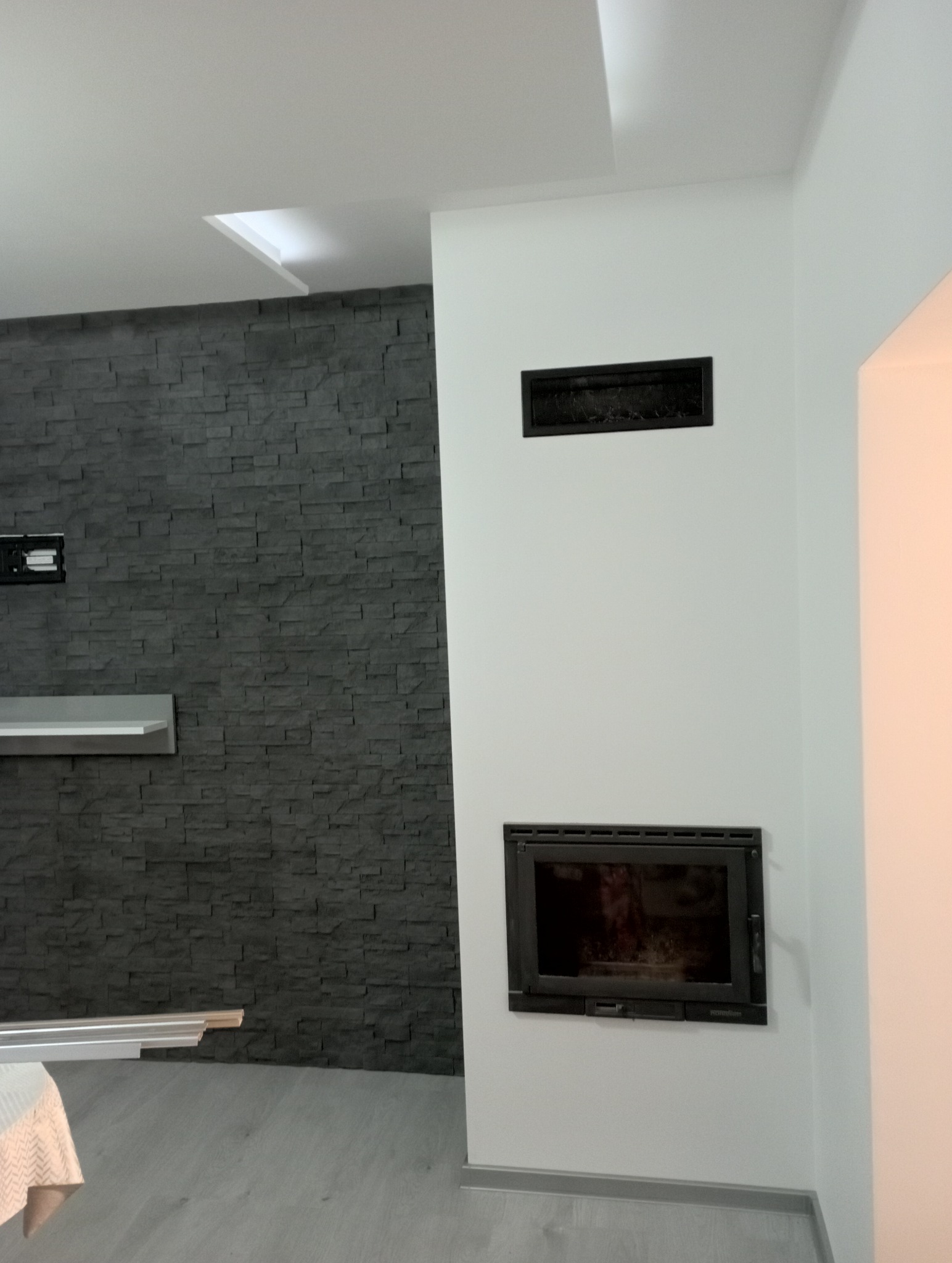 Nowoczesny salon z kominkiem, ściana wyłożona szarym kamieniem dekoracyjnym, widoczna półka i listwy przypodłogowe, biały sufit z wnęką.