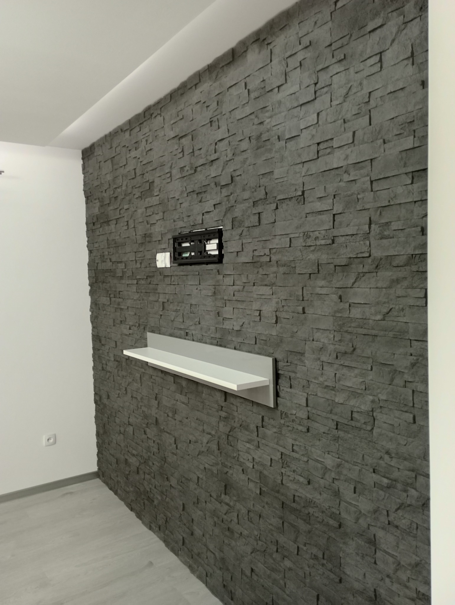 Ściana wykończona szarą okładziną kamienną, z zamontowanym uchwytem do telewizora i białą półką, widok z boku.