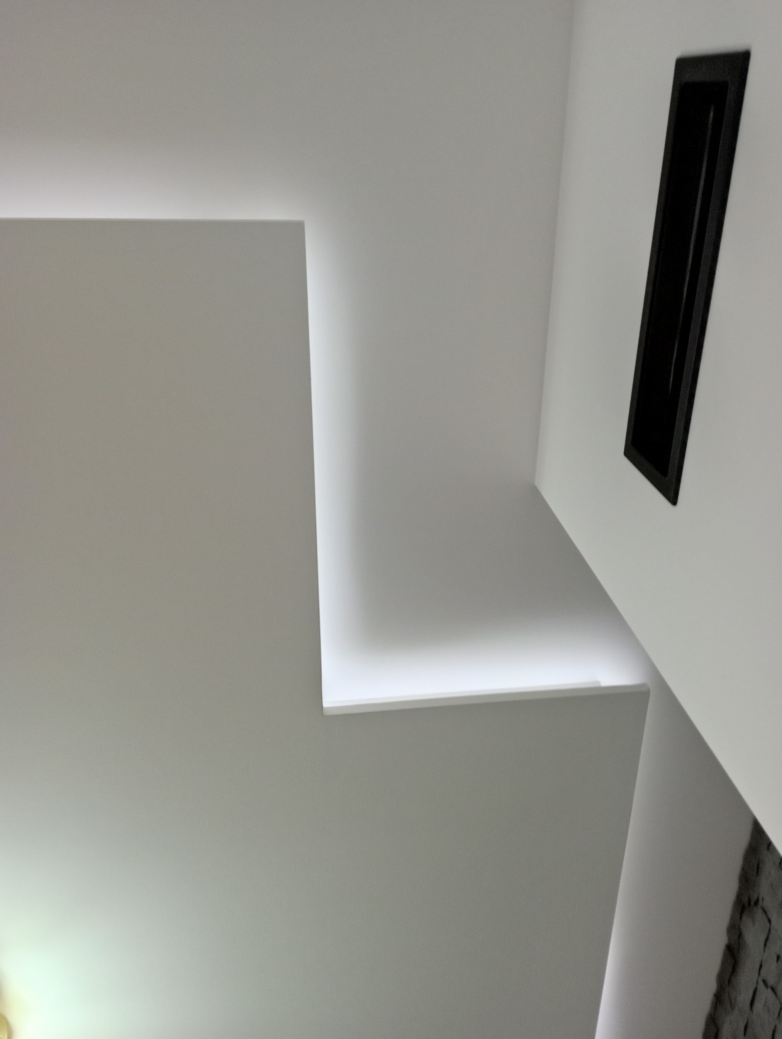 Nowoczesny, biały sufit z ukrytym oświetleniem LED, tworzącym geometryczne kształty, oraz czarny element dekoracyjny w ścianie.