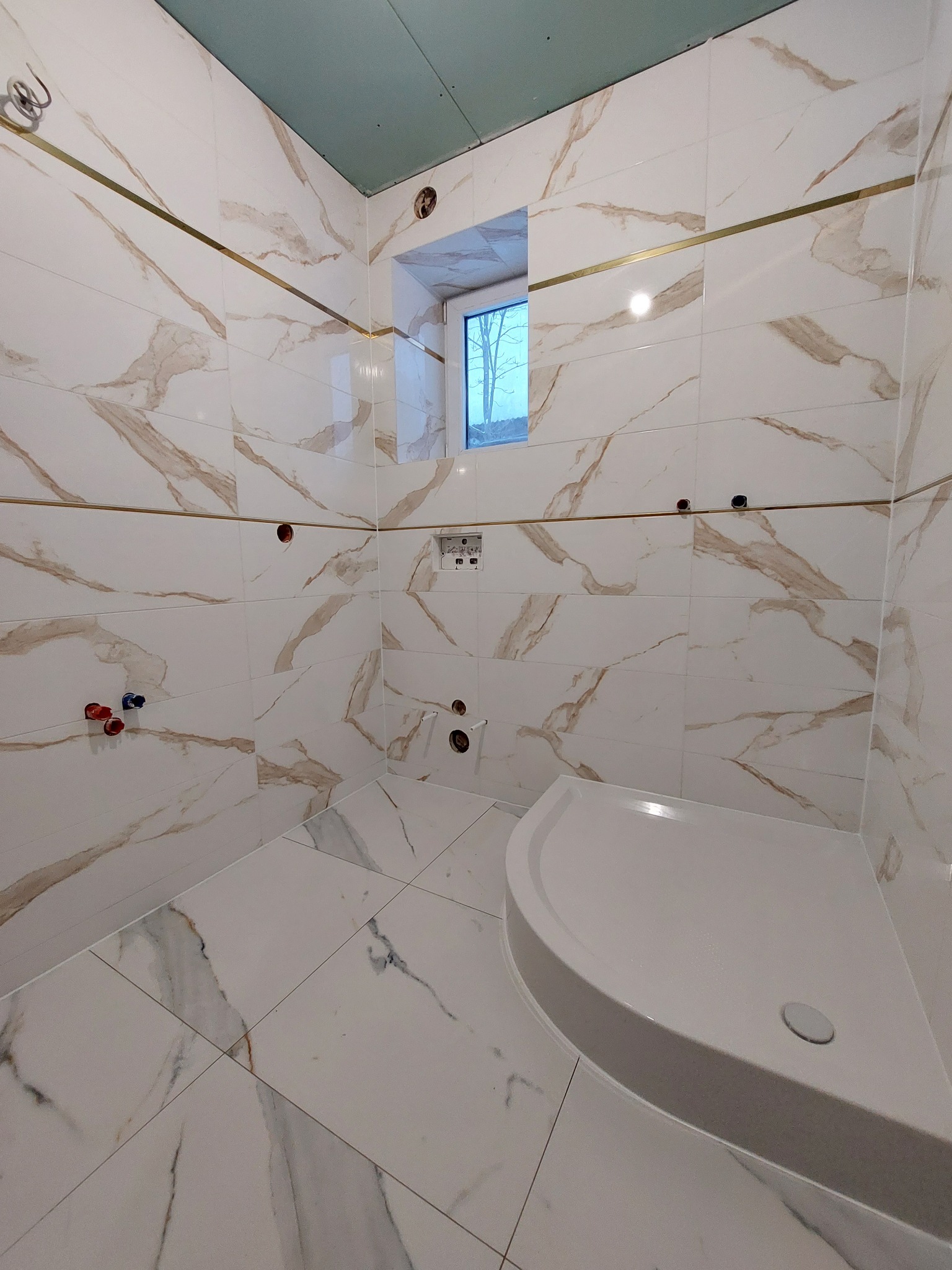 Wnętrze łazienki w trakcie prac wykończeniowych, widoczne białe płytki z marmurkowym wzorem i złotymi listwami, zamontowany brodzik, sufit z płyt gipsowo-kartonowych, przygotowane otwory na armaturę.