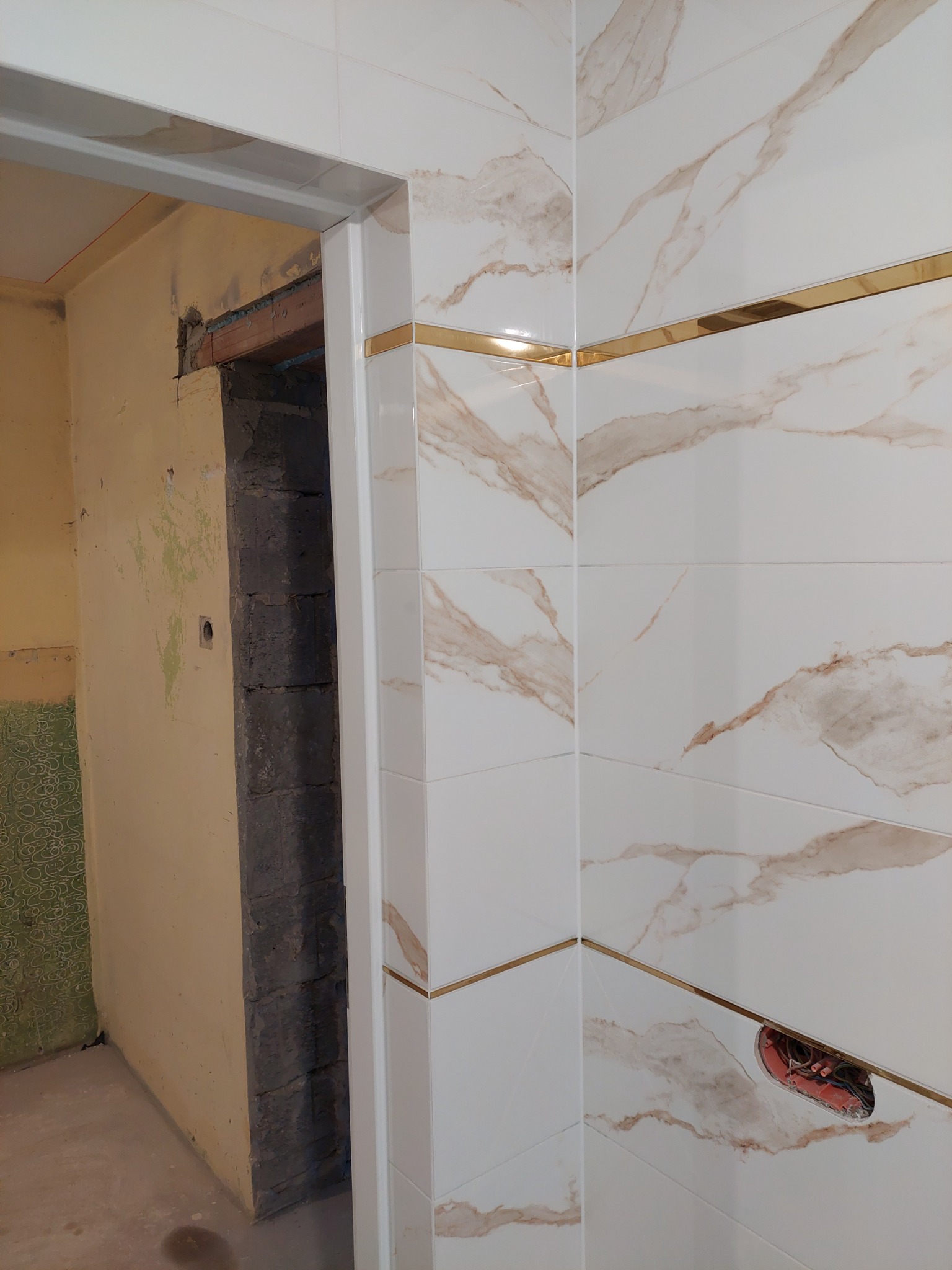 Fragment łazienki w trakcie remontu: ściana wyłożona białymi płytkami z marmurkowym wzorem i złotymi akcentami, widoczna puszka elektryczna i surowa ściana obok ościeżnicy.