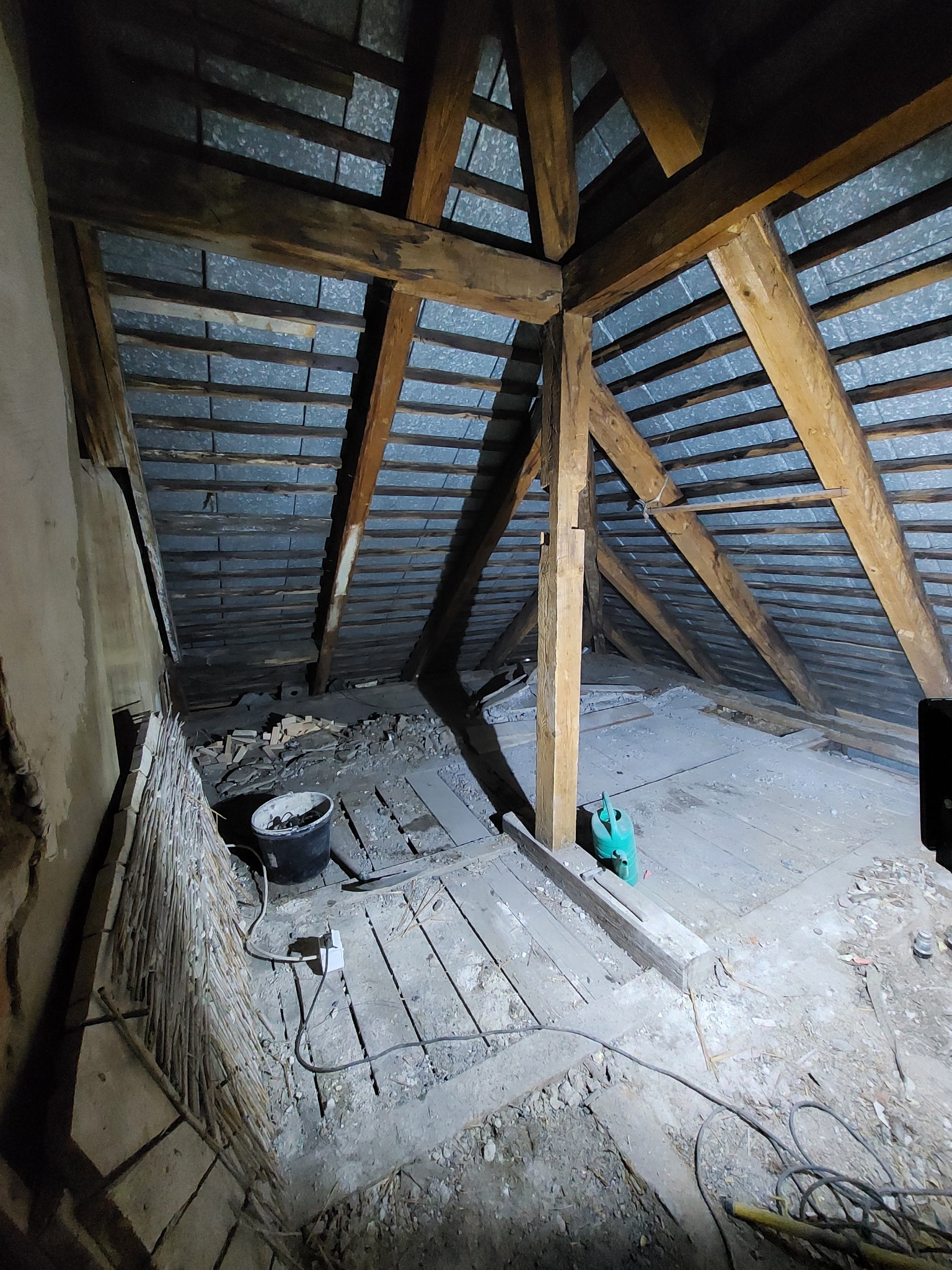 Poddasze w trakcie rozbiórki, widoczne drewniane belki konstrukcyjne, fragmenty cegieł, wiadro i zielony kanister, podłoga z desek częściowo zdemontowana.
