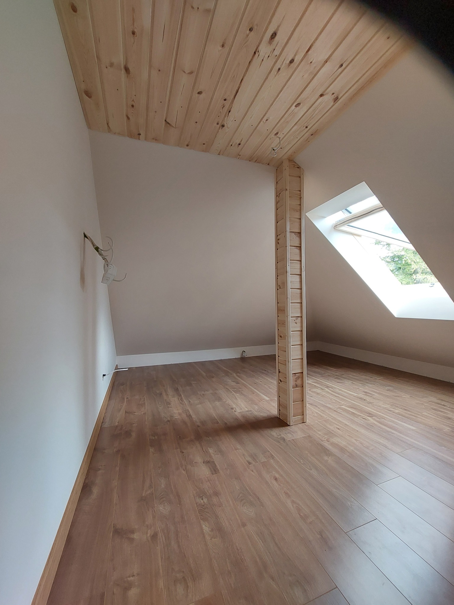 Poddasze z drewnianym wykończeniem sufitu i słupem obłożonym deskami, okno dachowe w skosie, brązowe panele podłogowe, ściany pomalowane na biało, widoczne kable na ścianie.