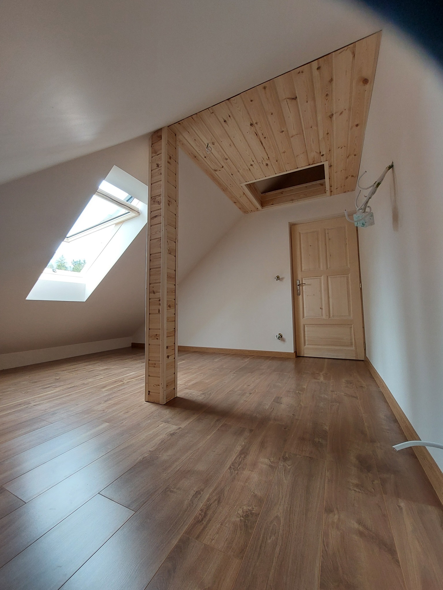 Poddasze po remoncie z drewnianą kolumną nośną, wykończonym sufitem z desek i panelami podłogowymi, widoczne okno dachowe i instalacja elektryczna.