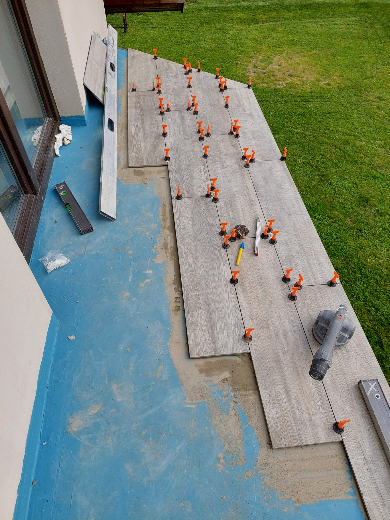 Układanie płytek na balkonie z użyciem systemu poziomowania, widoczne poziomice, ołówek i szlifierka kątowa.