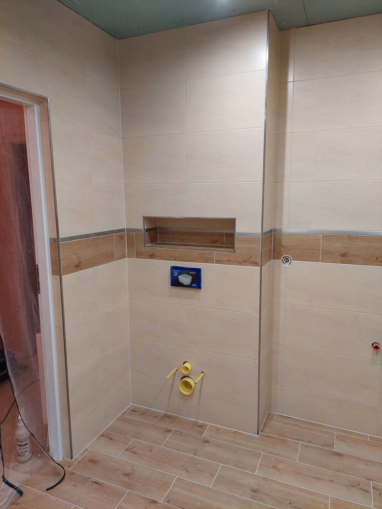 Wnętrze łazienki w trakcie prac wykończeniowych, widoczne beżowe płytki na ścianach z dekoracyjnym pasem imitującym drewno, płytki podłogowe w odcieniach drewna, zamontowany stelaż podtynkowy WC...