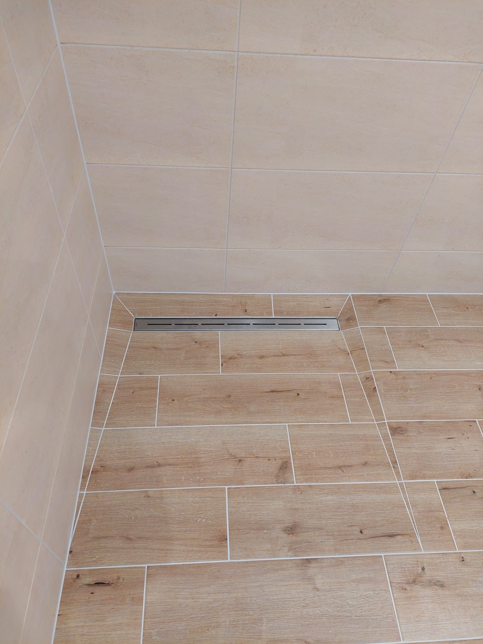 Beżowe płytki ścienne i podłogowe imitujące drewno z odpływem liniowym w rogu kabiny prysznicowej.