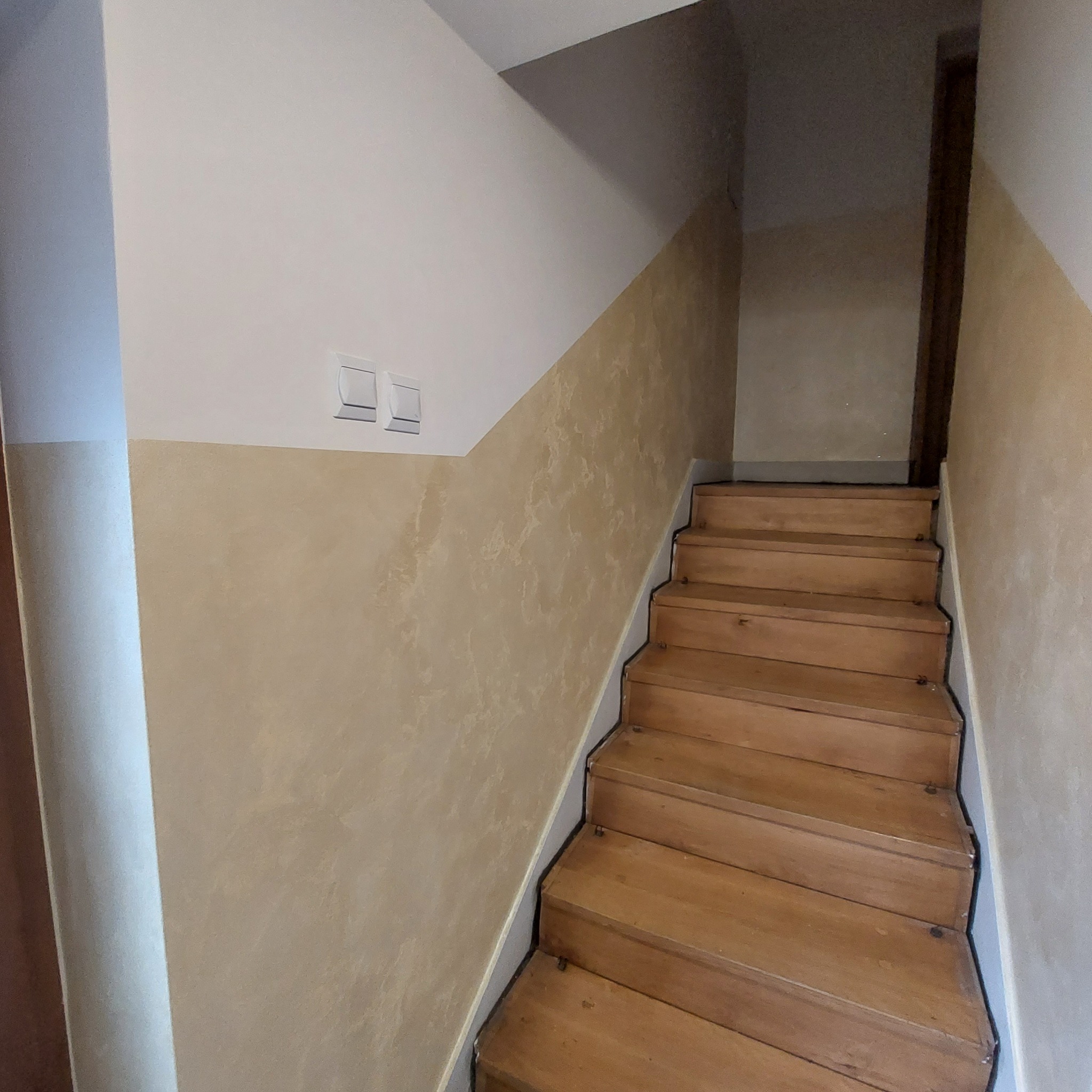 Klatka schodowa z drewnianymi schodami, ściany pomalowane na beżowo z efektem dekoracyjnym, widoczne dwa włączniki światła na górze ściany.