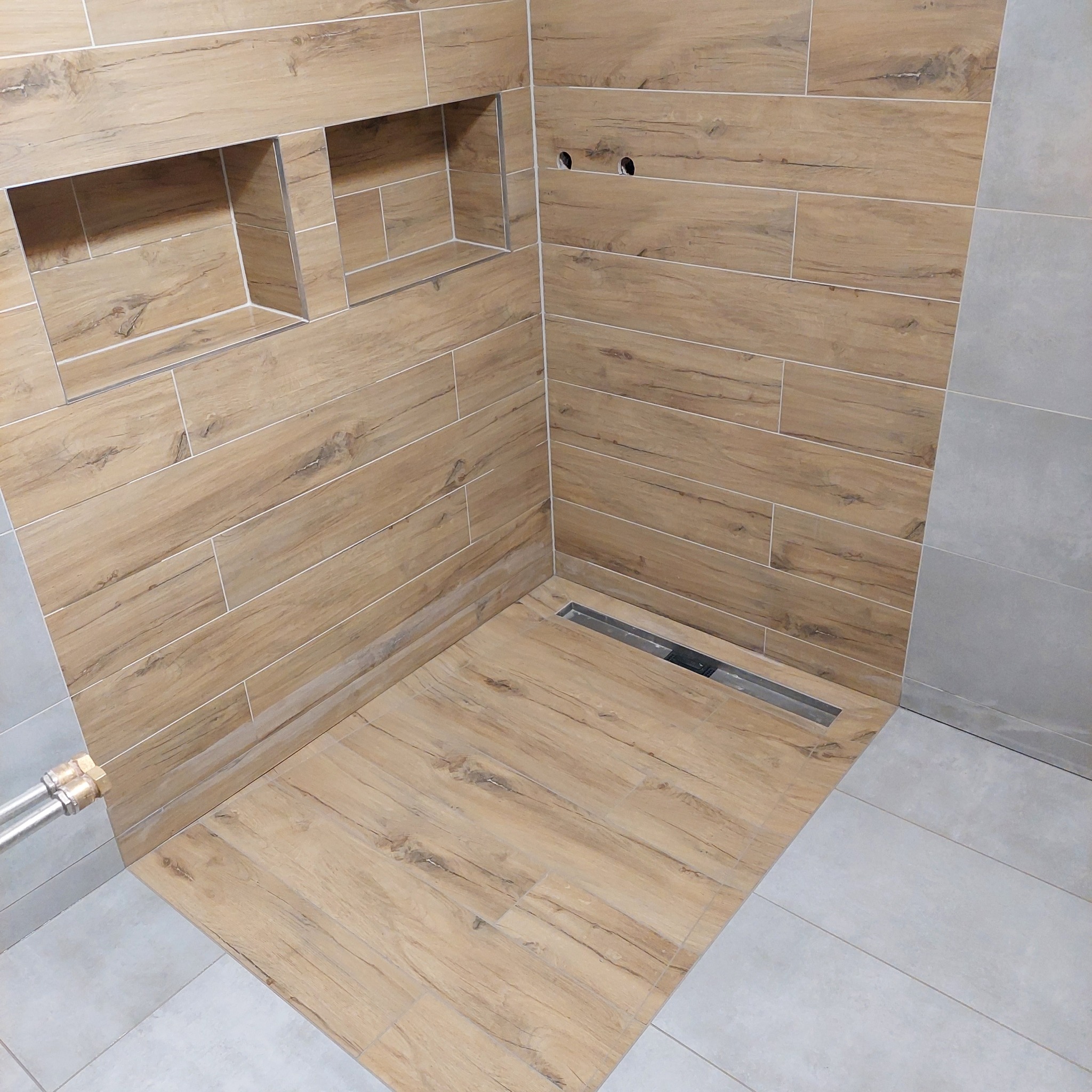 Nowoczesna kabina prysznicowa wyłożona płytkami imitującymi drewno, z wbudowanymi półkami i odpływem liniowym, połączona z szarymi płytkami na podłodze i ścianie.