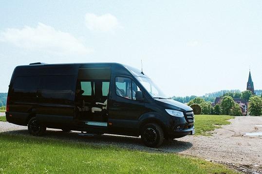 Czarny van Mercedes-Benz Sprinter z otwartymi drzwiami przesuwnymi, zaparkowany na trawiastym wzgórzu z widokiem na kościół z wieżą w tle.