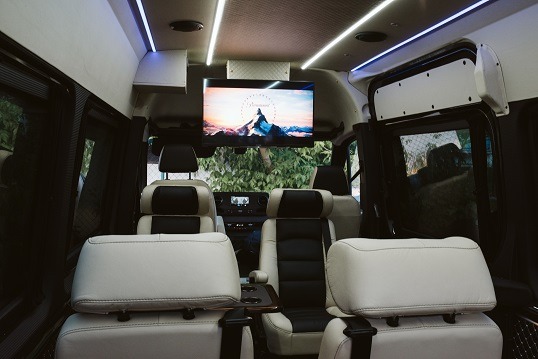 Wnętrze luksusowego busa z kremowymi skórzanymi fotelami, drewnianym stolikiem i ekranem wyświetlającym logo Paramount Pictures.