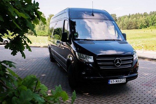 Czarny Mercedes-Benz Sprinter z polską tablicą rejestracyjną zaparkowany na brukowanym placu, częściowo zasłonięty przez gałęzie drzewa.