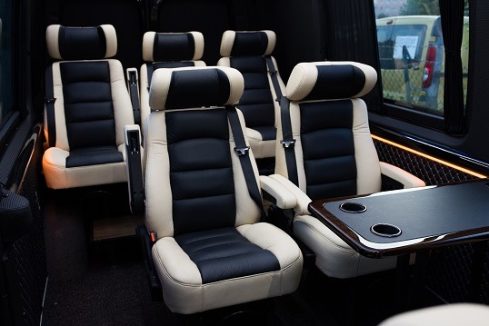 Wnętrze luksusowego busa z beżowo-czarnymi skórzanymi fotelami i rozkładanym stolikiem z uchwytami na napoje.