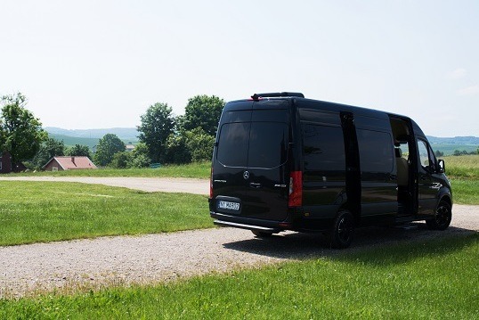 Czarny, luksusowy van Mercedes-Benz Sprinter z otwartymi drzwiami, stojący na żwirowej drodze w otoczeniu zielonych łąk i drzew, z widocznym domem z czerwoną dachówką w tle.