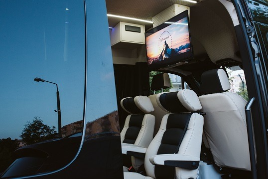 Wnętrze luksusowego busa do przewozu osób z jasną skórzaną tapicerką, zamontowanym telewizorem z logo Paramount i oświetleniem LED.