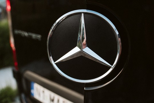 Chromowany znak Mercedesa na czarnym samochodzie dostawczym, widoczne fragmenty tablicy rejestracyjnej i tylnej lampy.