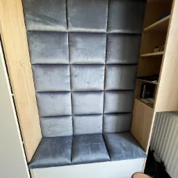 Wnęka z siedziskiem obitym szarymi, kwadratowymi panelami tapicerowanymi, otoczona drewnianymi regałami i szafkami w jasnym odcieniu drewna. Na podłodze widoczne obuwie i narzędzia malarskie.