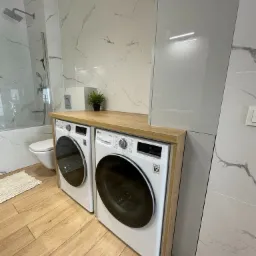 Pralka i suszarka LG zabudowane pod drewnianym blatem w łazience z marmurowymi płytkami, obok kabina prysznicowa.