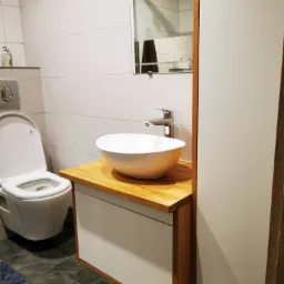 Nowoczesna łazienka z umywalką nablatową w kształcie misy na drewnianym blacie, szafka pod umywalką z białymi frontami i drewnianymi bokami, obok wysoka szafa, w tle toaleta i białe kafelki.