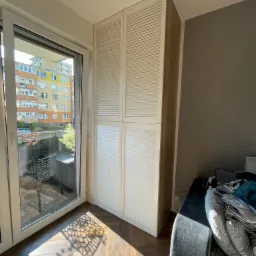 Biała szafa z żaluzjowymi drzwiami, wbudowana obok okna balkonowego, z widocznym parkietem w jodełkę i fragmentem sofy.