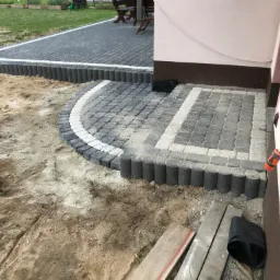 Fragment układania szarej kostki brukowej z białymi akcentami i obrzeżami z elementów cylindrycznych, widoczne przygotowane podłoże z piasku i zapas kostki.