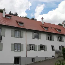 Budynek z nowym, czerwonym dachem ceramicznym, białymi ścianami i szarymi okiennicami, widok z dołu na tle pochmurnego nieba.