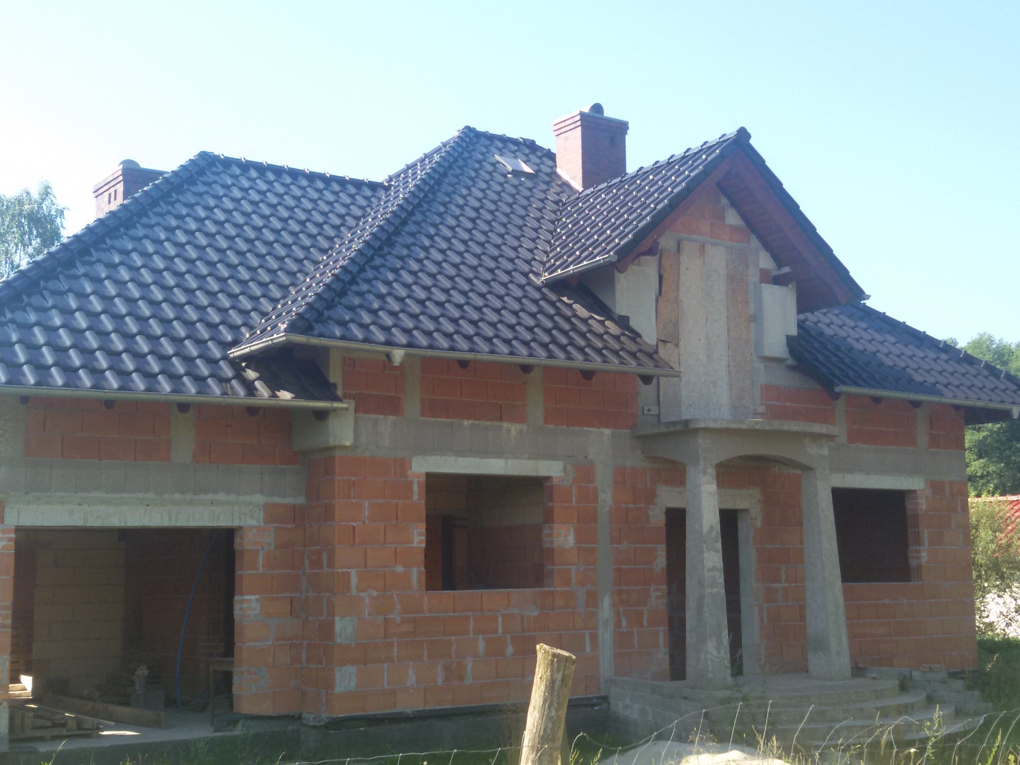 Dom w trakcie budowy z nowym, ciemnym dachem z dachówki ceramicznej, widoczna surowa cegła i niezabudowane otwory okienne.