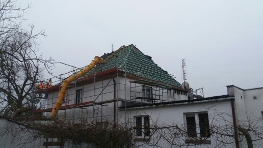 Prace na dachu dwukondygnacyjnego budynku z zieloną konstrukcją nośną, żółta rura zsypowa i rusztowania w pochmurny dzień.