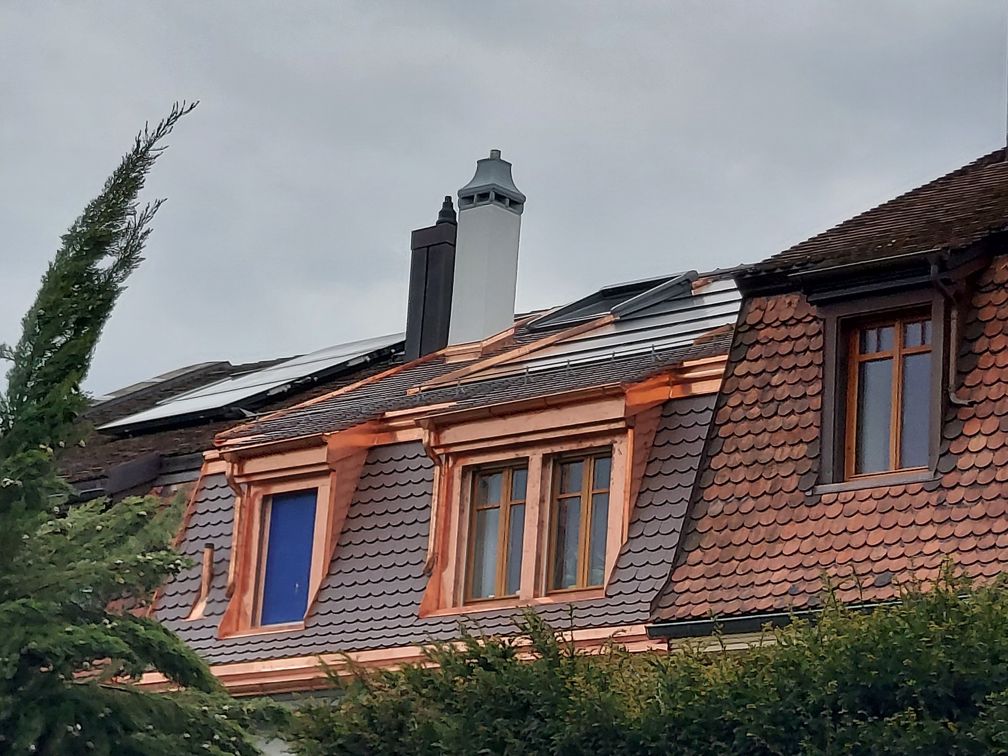 Dach z lukarnami, pokryty dachówką karpiówką i obróbkami blacharskimi z miedzi, widoczne kominy i panele słoneczne.