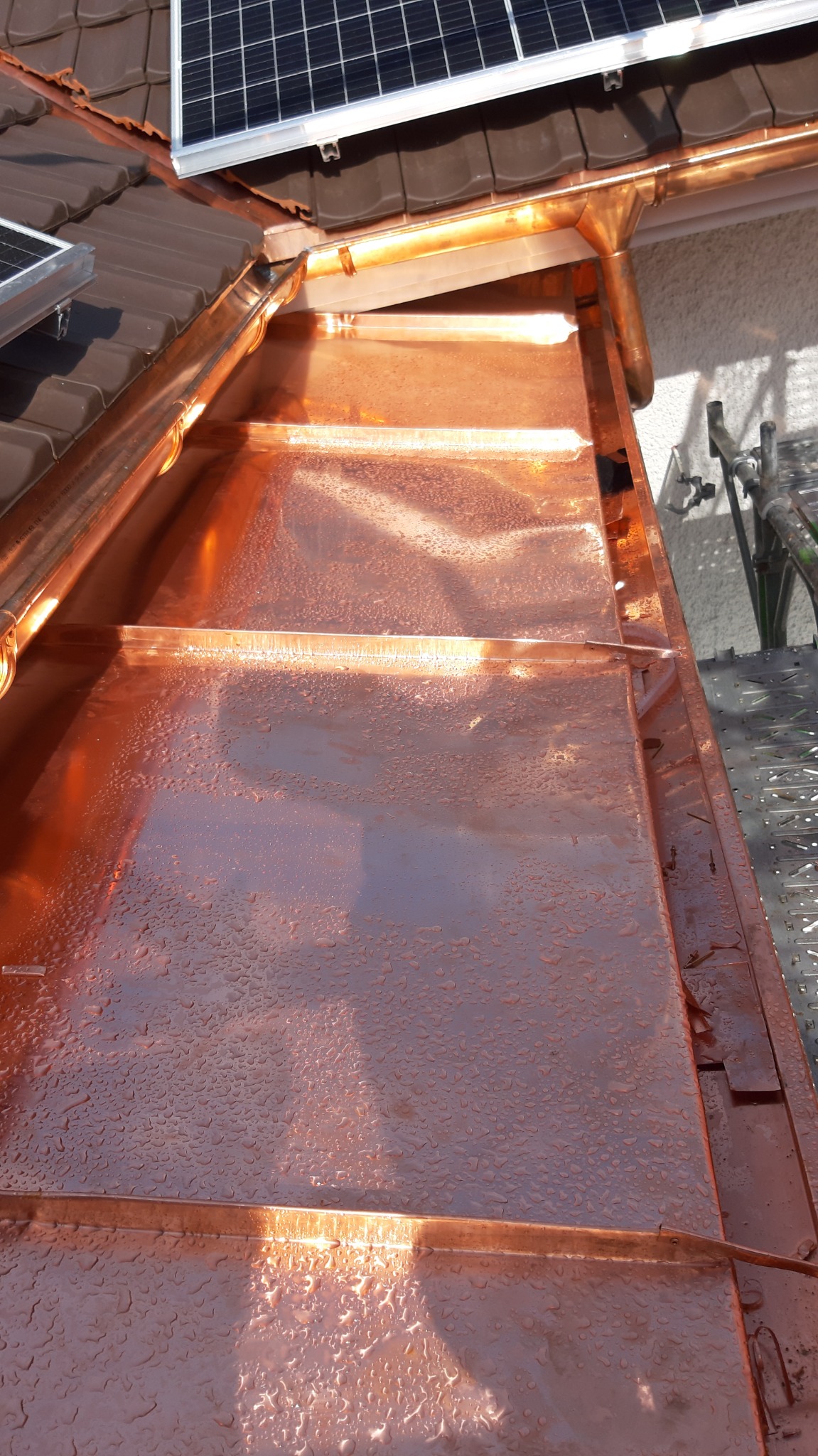 Miedziane orynnowanie dachu z panelami słonecznymi, pokryte kroplami wody, widoczne z bliska.